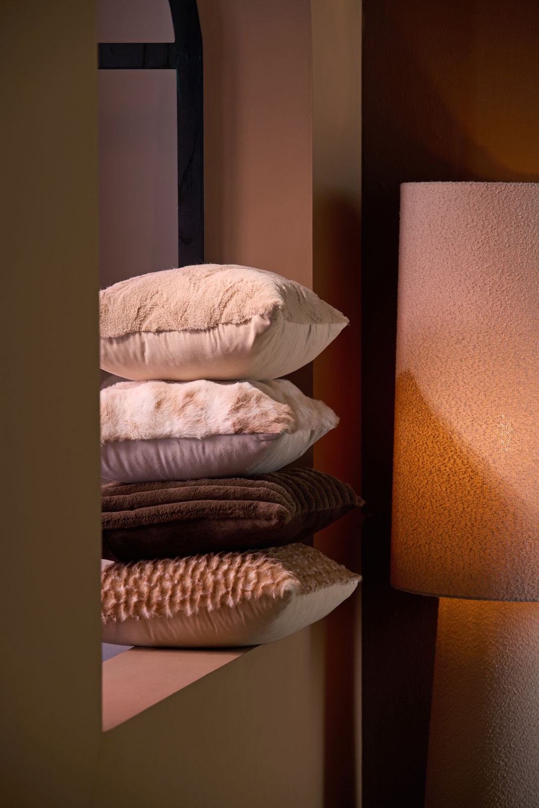 Ein Stapel von vier Kissen in verschiedenen Brauntönen, die auf einem Regal neben einer Lampe liegen. Die Kissen haben unterschiedliche Texturen und Muster. Perspektive von der Seite.