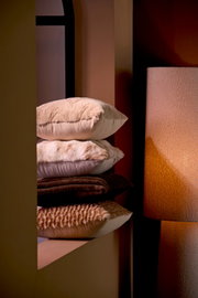 Ein Stapel von vier Kissen in verschiedenen Brauntönen, die auf einem Regal neben einer Lampe liegen. Die Kissen haben unterschiedliche Texturen und Muster. Perspektive von der Seite.
