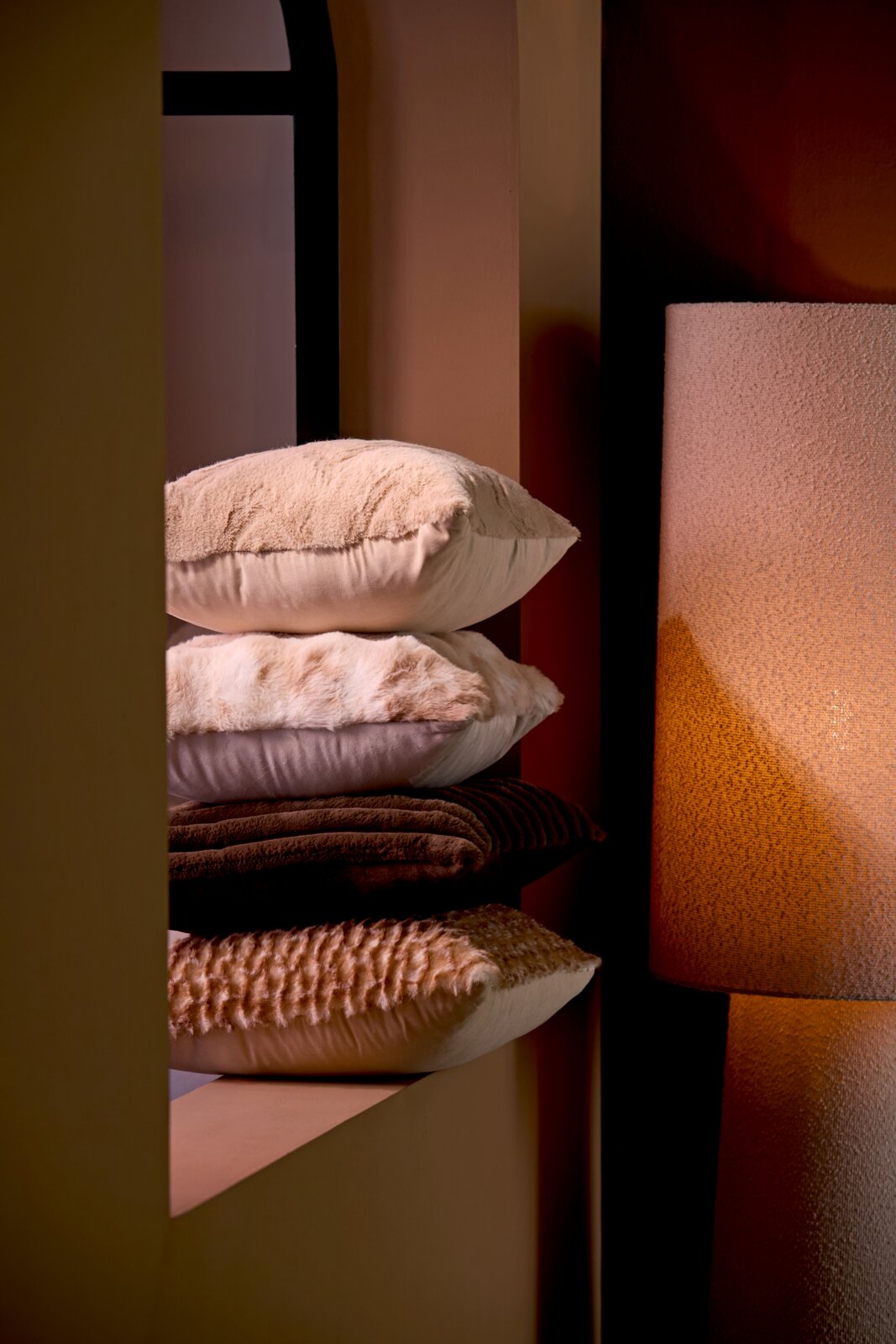 Ein Stapel von vier Kissen in verschiedenen Brauntönen, die auf einem Regal neben einer Lampe liegen. Die Kissen haben unterschiedliche Texturen und Muster. Perspektive von der Seite.