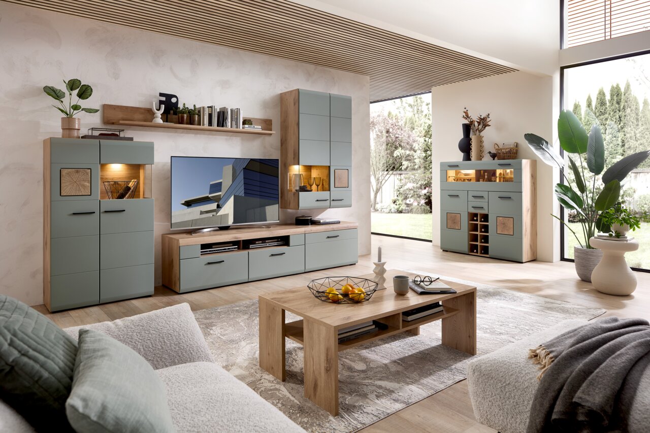 Moderne Wohnwand in einem hellen Wohnzimmer mit großen Fenstern, bestehend aus mehreren Schränken und einem TV-Board in Holzoptik und grauen Fronten, seitliche Perspektive.