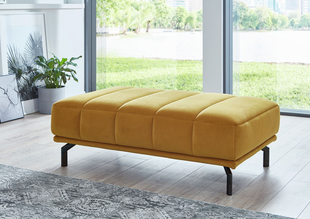 Gelber XXL-Hocker in modernem Wohnzimmer, seitliche Perspektive mit Blick auf große Fenster und grüne Landschaft im Hintergrund.