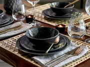 Schwarz-glänzendes Geschirr-Set mit Schüssel und Teller auf geflochtener Tischmatte, daneben Glas und goldfarbenes Besteck; perspektivisch in schräger Draufsicht von vorne fotografiert.