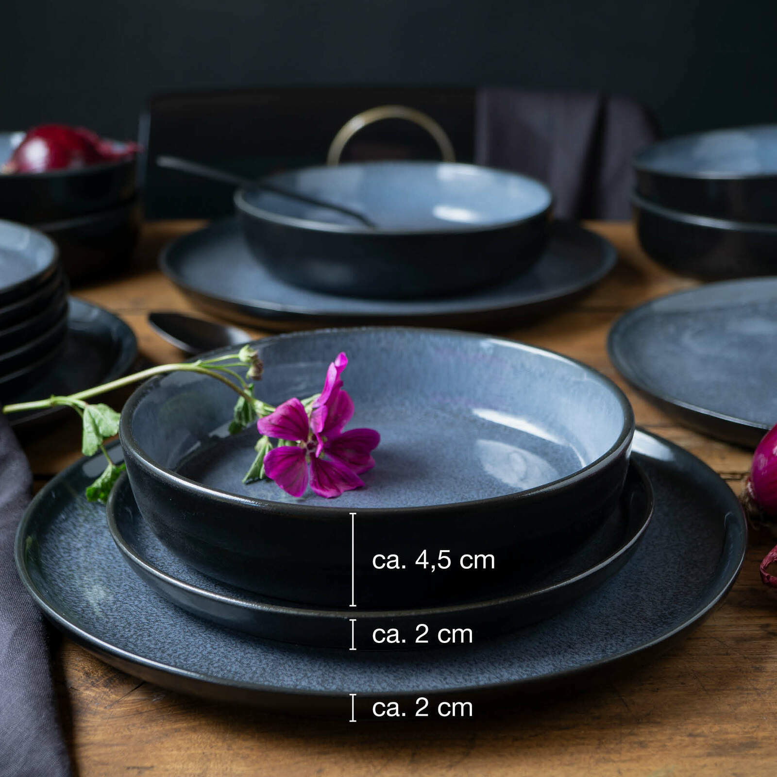 Ein elegantes, blaues 18-teiliges Tafelservice, bestehend aus Tellern und Schalen, auf einem Holztisch arrangiert. Im Vordergrund eine Schale mit einer violetten Blume, seitliche Perspektive.