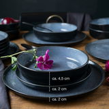 Ein elegantes, blaues 18-teiliges Tafelservice, bestehend aus Tellern und Schalen, auf einem Holztisch arrangiert. Im Vordergrund eine Schale mit einer violetten Blume, seitliche Perspektive.
