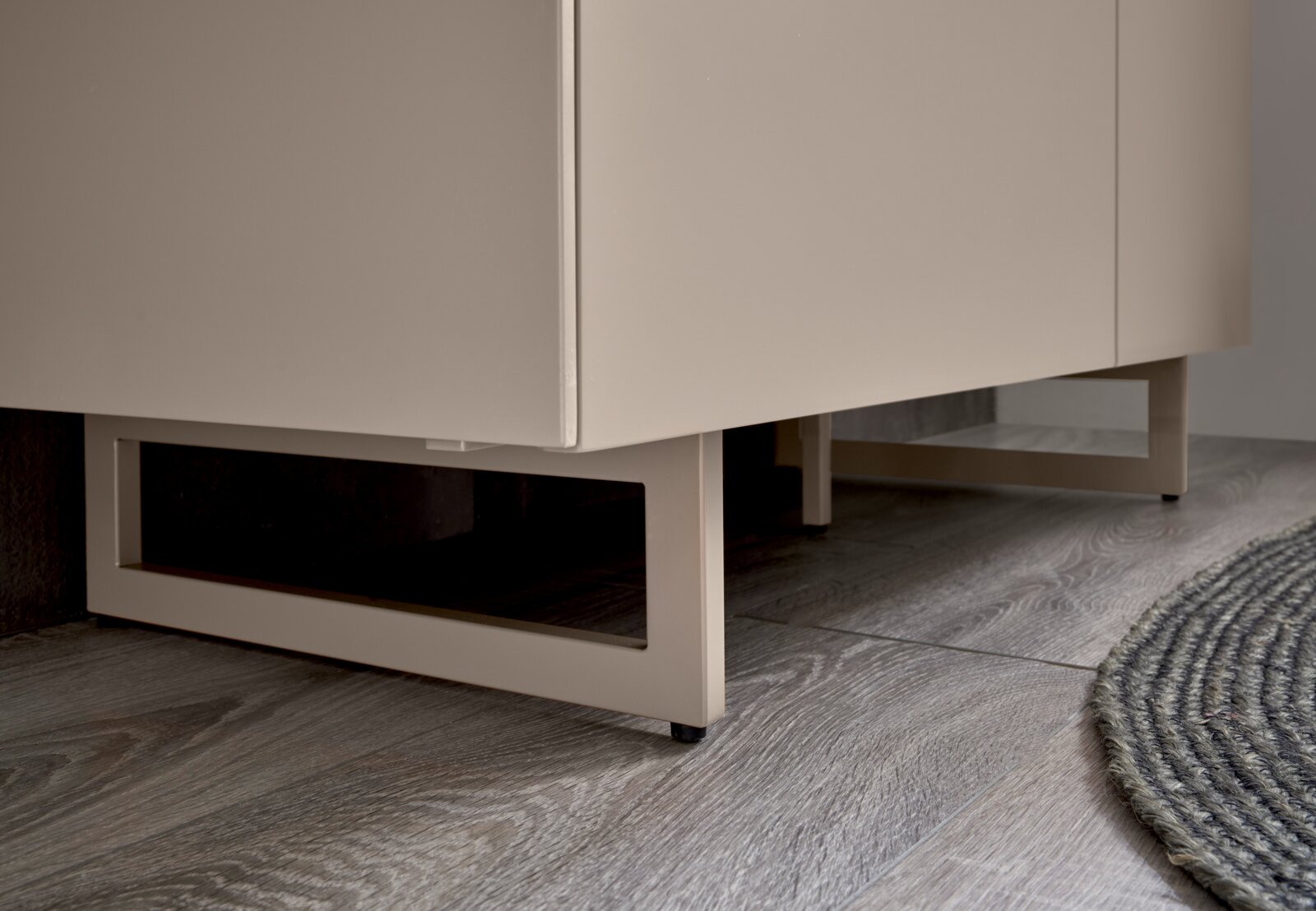 Detailaufnahme aus niedriger Perspektive: weißer Badezimmerschrank/Waschtischunterschrank mit offenem Metallgestell und schwarzen Füßen auf grauem Holzfußboden neben rundem Teppich