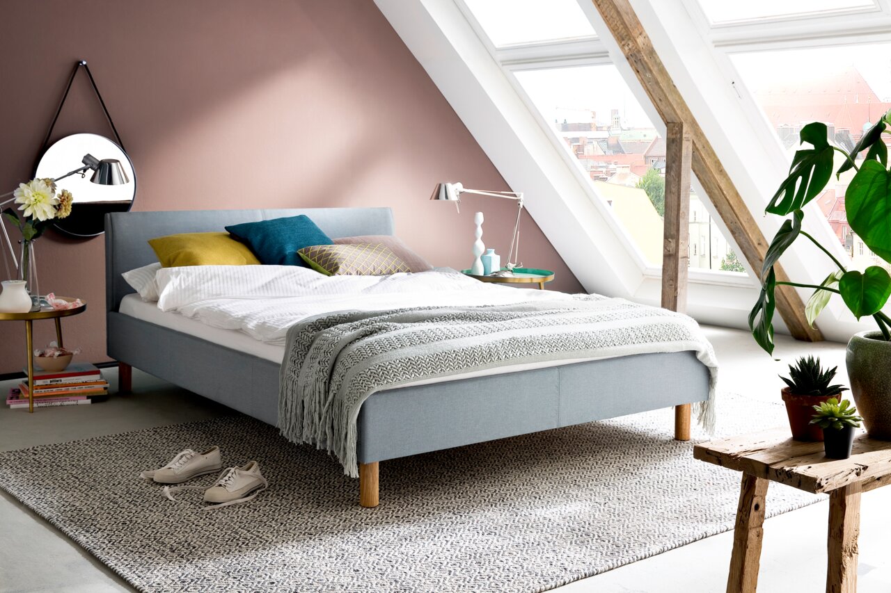 Modernes Polsterbett in einem stilvollen Dachgeschosszimmer mit schrägen Fenstern, seitliche Perspektive.
