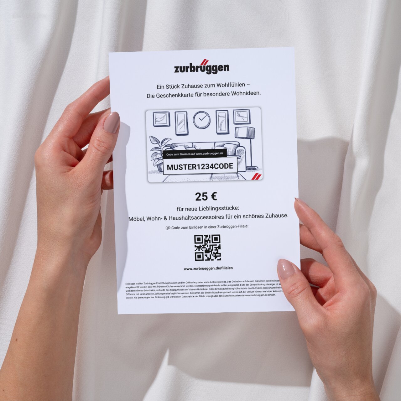 Zurbrüggen Digitaler Geschenkgutschein zum Ausdrucken Zwei Hände halten eine Zurbrüggen-Geschenkkarte im Wert von 25 Euro, fotografiert aus der Vogelperspektive. Die Karte zeigt ein Wohnzimmerdesign und enthält einen QR-Code zur Einlösung.