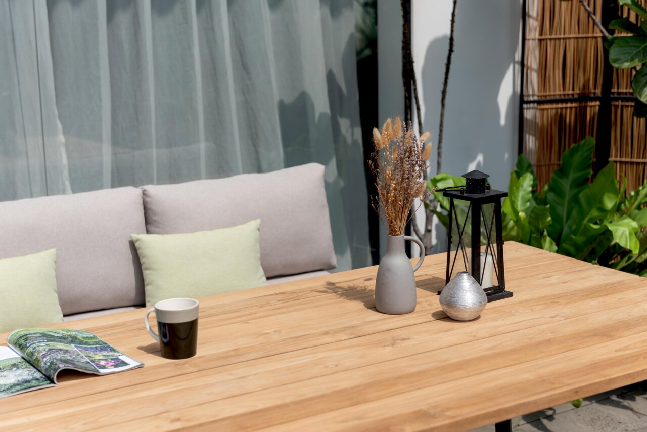 Gartentisch aus Holz mit Dekorationen, darunter eine Vase mit Trockenblumen, eine Laterne und eine Tasse, im Freien auf einer Terrasse mit Sitzbank im Hintergrund, seitliche Perspektive.