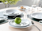 Modern gedeckter Esstisch mit weißem Geschirr, Besteck und Wassergläsern; im Vordergrund liegt ein grüner Apfel auf einem Teller – aufgenommen aus leicht erhöhter, schräger Frontperspektive.