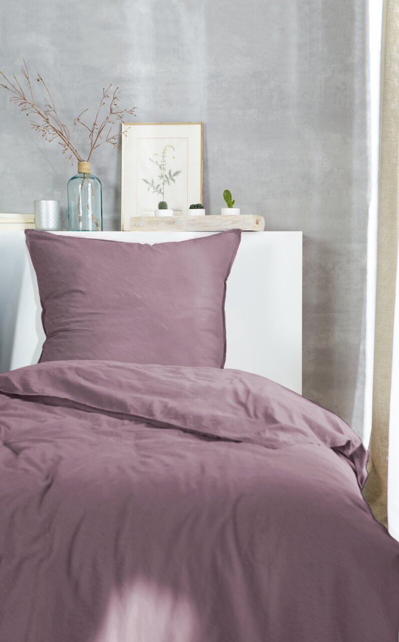 Lila Bettwäsche-Set auf einem Bett, frontal fotografiert, mit dekorativen Elementen im Hintergrund.