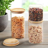 Drei Aufbewahrungsgläser mit Bambusdeckeln, gefüllt mit Nudeln, Müsli und getrockneten Früchten, auf einem Holztisch in einer hellen Umgebung.