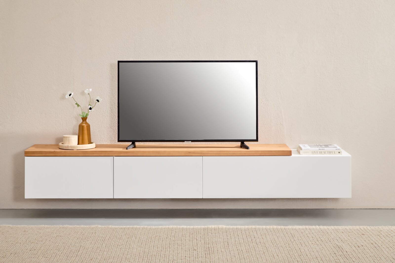Modernes, schwebendes TV-Lowboard in Weiß mit Holzoberfläche, darauf ein Flachbildfernseher sowie Vase und Bücher Frontalansicht auf Augenhöhe.