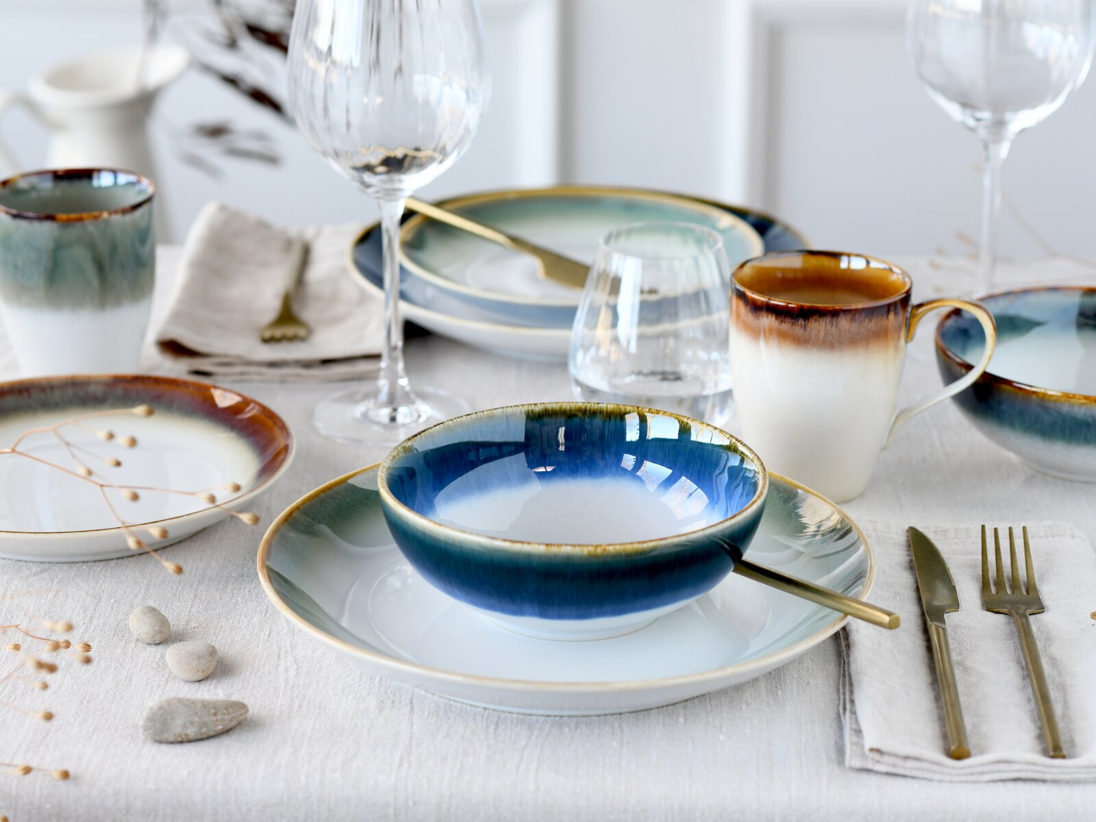 Schick gedeckter Esstisch mit blau-weiß glasiertem Keramikgeschirr, Tasse und Schalen, goldfarbenem Besteck und Weingläsern, aus leicht erhöhter Frontperspektive