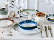 Schick gedeckter Esstisch mit blau-weiß glasiertem Keramikgeschirr, Tasse und Schalen, goldfarbenem Besteck und Weingläsern, aus leicht erhöhter Frontperspektive