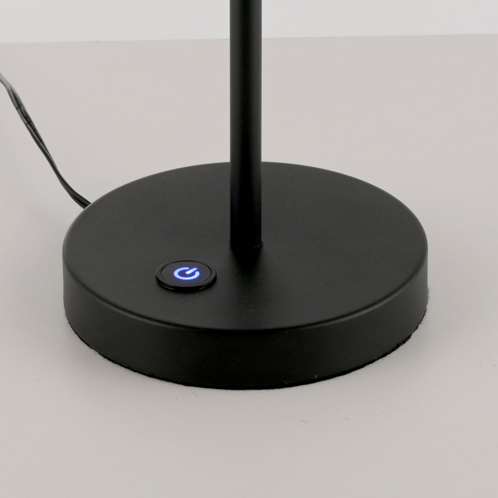Schwarzer runder Lampenfuß einer Tischlampe mit blau beleuchtetem Ein-/Aus-Touchknopf und sichtbarem Kabel, Nahaufnahme in leichter Draufsicht von vorne