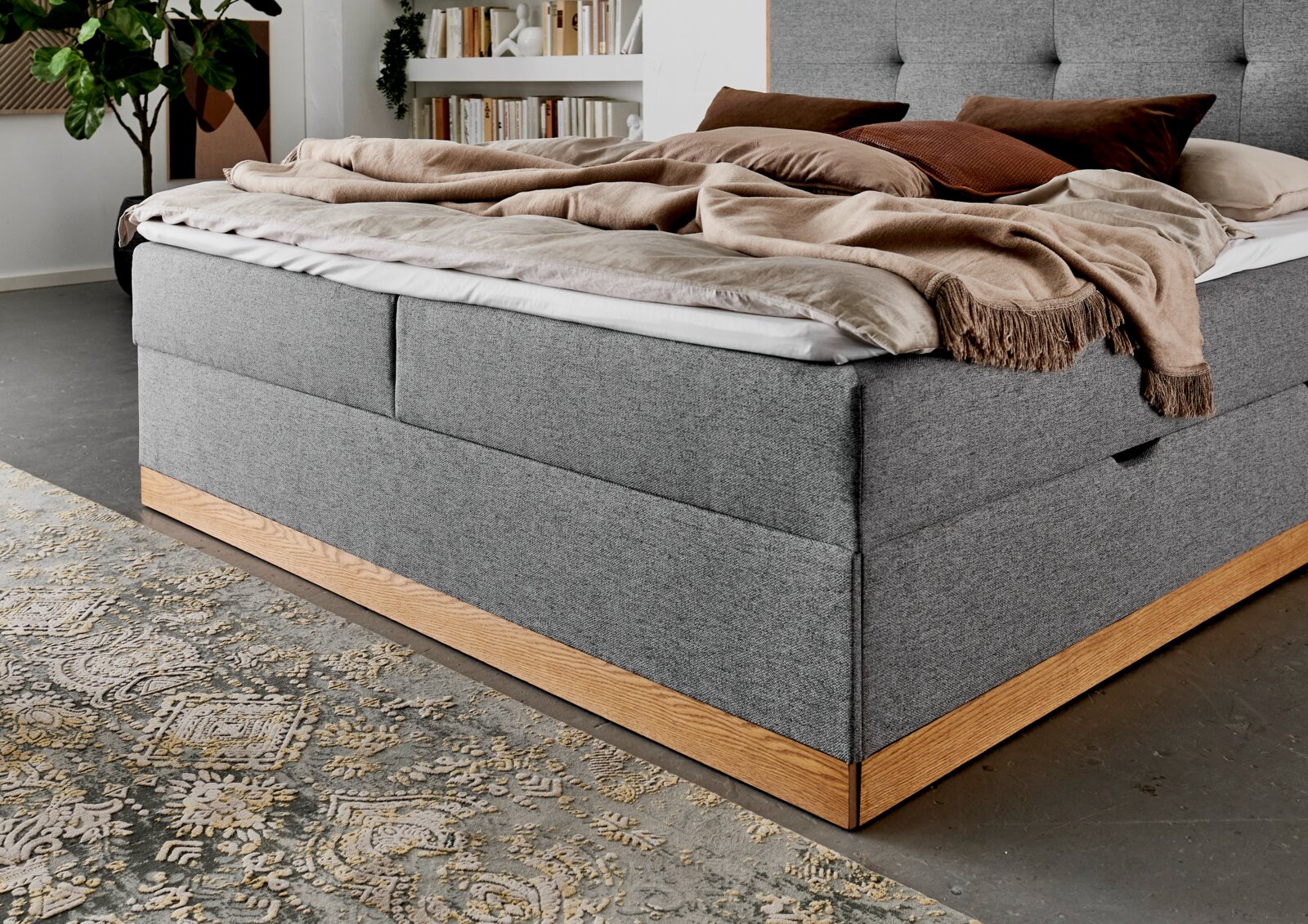 Grijs boxbed met houten frame, gezien vanaf de zijkant, versierd met kussens en dekens in een moderne slaapkamer.
