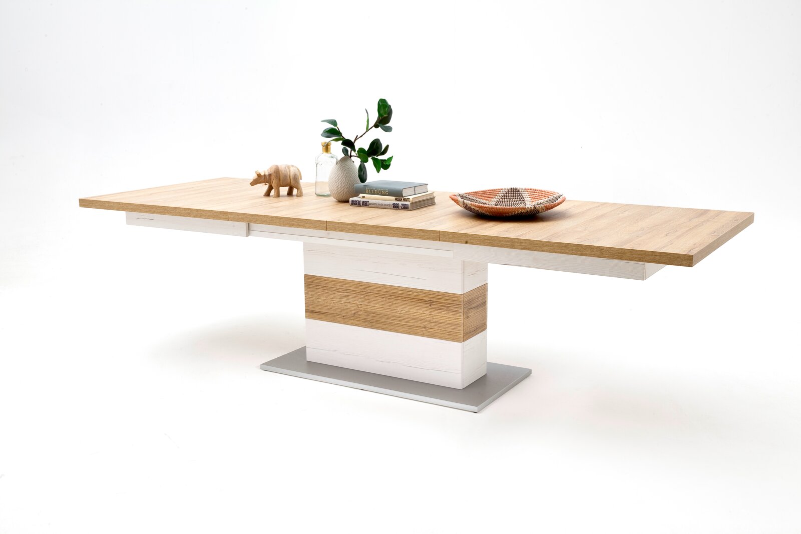 Moderner ausziehbarer Esstisch mit heller Holzplatte und weißem Mittelfuß auf grauer Bodenplatte, dekoriert mit Vase, Büchern und Schale, in leicht schräger Frontansicht vor weißem Hintergrund.