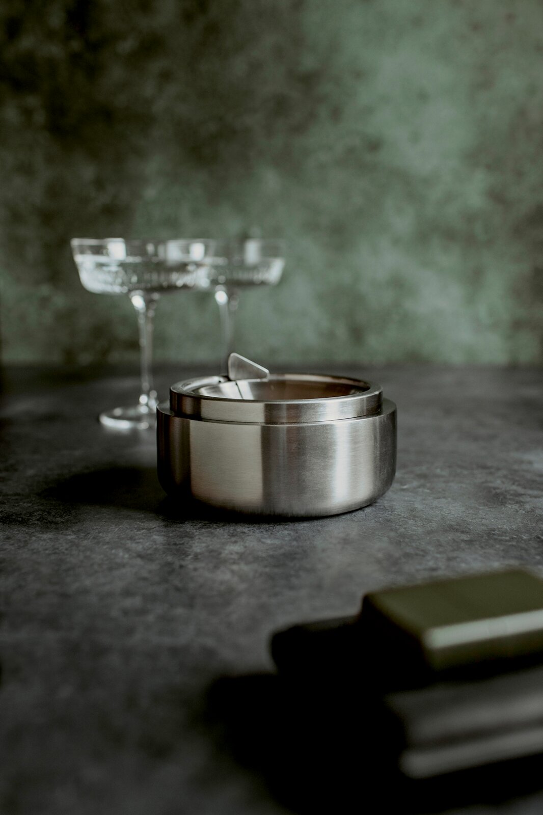 Edelstahl-Aschenbecher mit Deckel auf dunkler Steinoberfläche, leicht von vorne in niedriger Perspektive fotografiert im Hintergrund unscharfe Cocktailgläser.