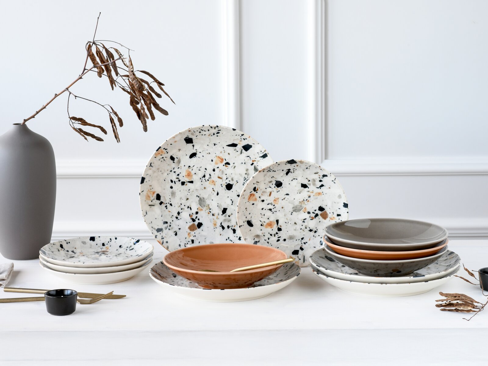 Modernes Geschirr-Set mit gesprenkelten Terrazzo-Tellern und Schalen in Weiß, Grau und Terrakotta, auf weißem Tisch mit Vase und Besteck, frontal in leichter Schrägansicht fotografiert.