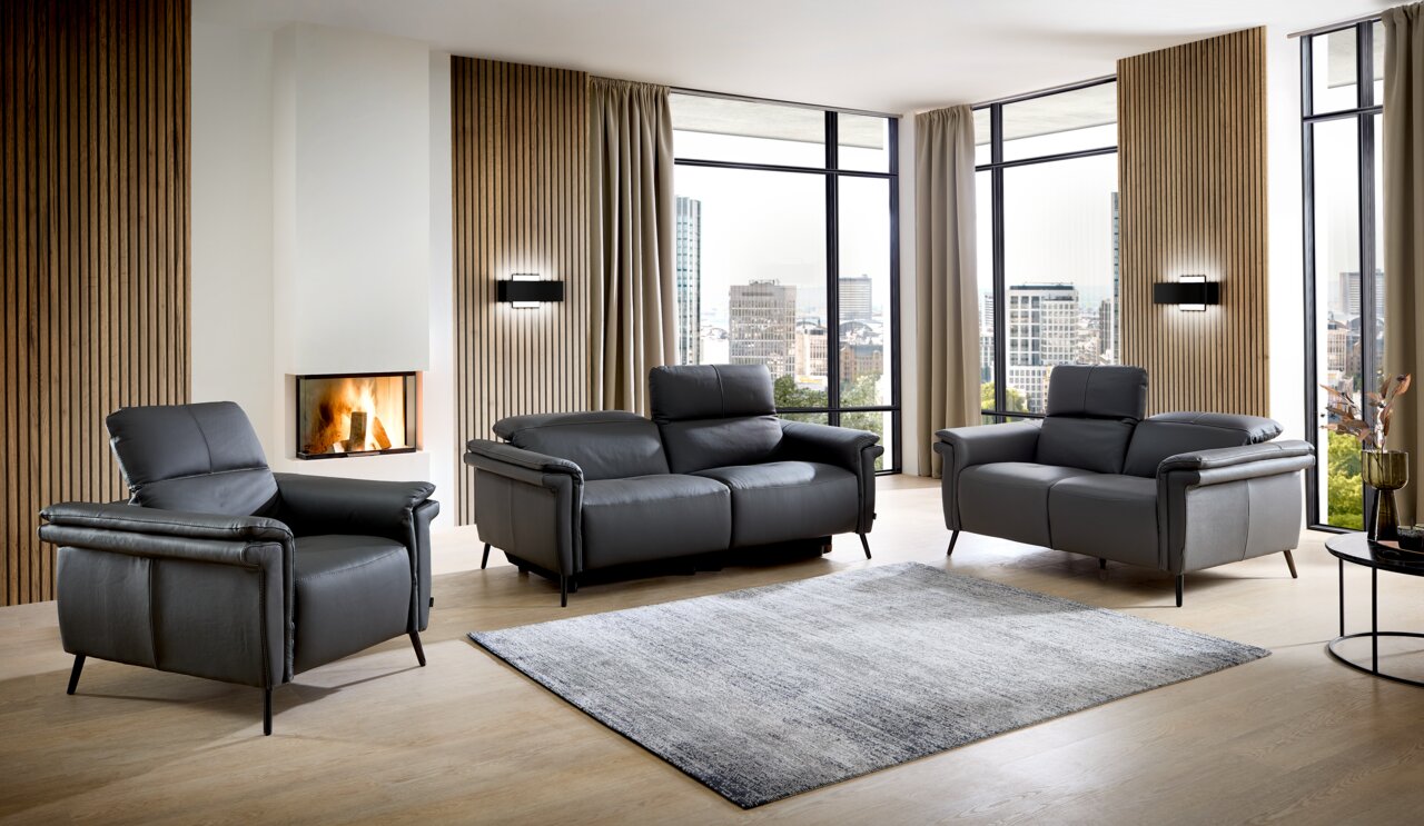 Wohnzimmer mit modernem, grauem Ledersofa-Set bestehend aus einem Dreisitzer, Zweisitzer und Sessel, vor einem Kamin und großen Fenstern mit Stadtblick, aus frontaler Perspektive.