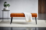 Sitzbank aus braunem Leder mit schwarzen Beinen, frontal fotografiert, dekoriert mit einer weißen Decke, neben einem kleinen Tisch mit Pflanze.
