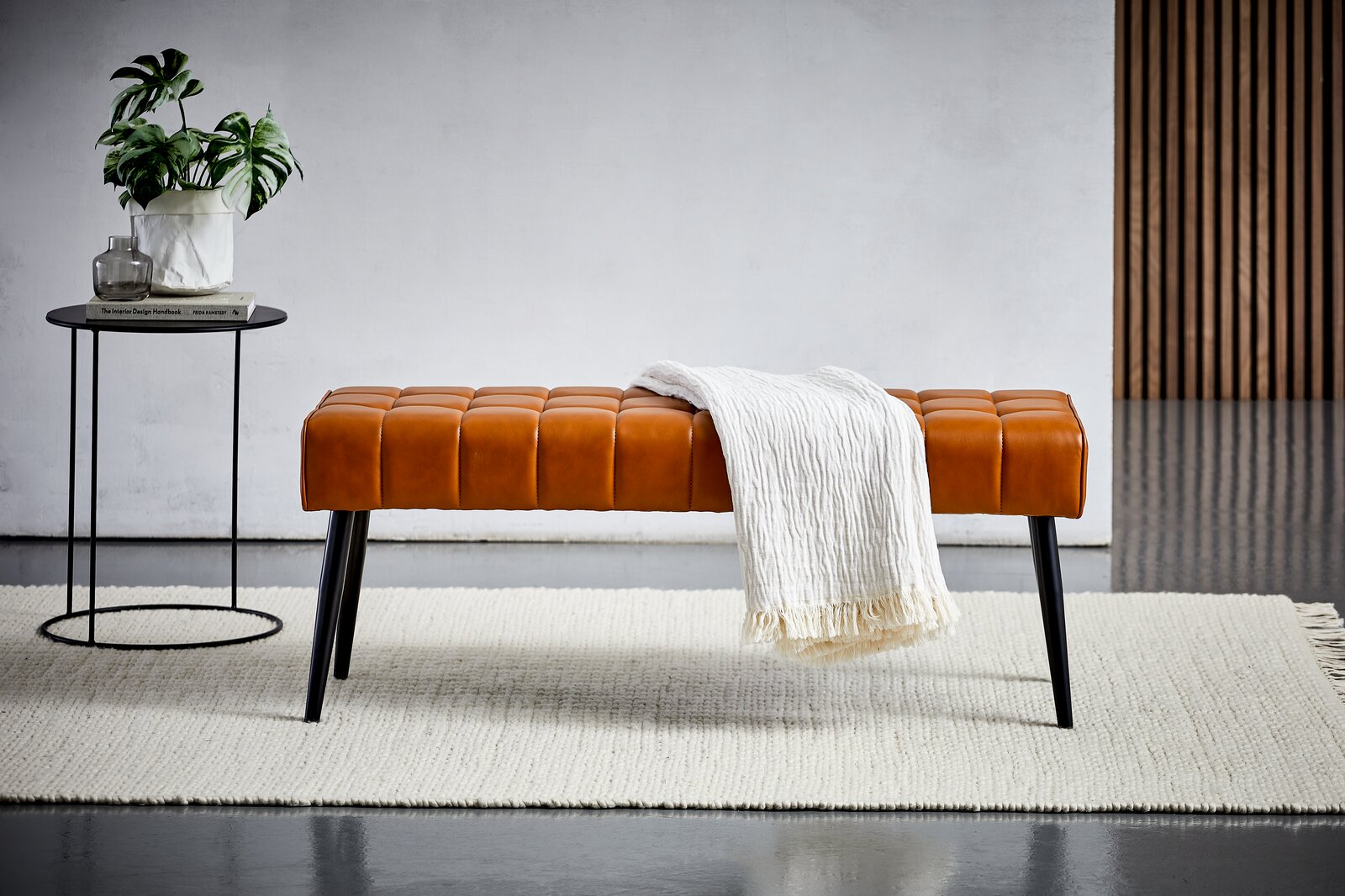 Sitzbank aus braunem Leder mit schwarzen Beinen, frontal fotografiert, dekoriert mit einer weißen Decke, neben einem kleinen Tisch mit Pflanze.