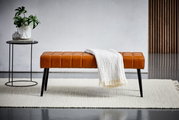 Sitzbank aus braunem Leder mit schwarzen Beinen, frontal fotografiert, dekoriert mit einer weißen Decke, neben einem kleinen Tisch mit Pflanze.