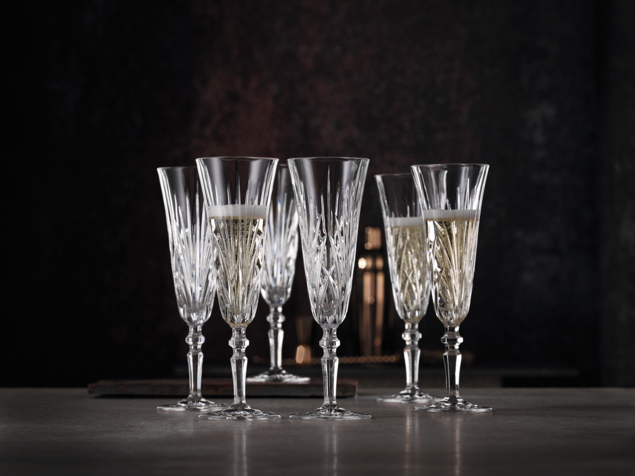 Nachtmann Sektspitz PALAIS Sechs elegante Sektgläser aus Kristall mit feinem Schliff, teilweise gefüllt mit Sekt, auf einem Tisch vor dunklem Hintergrund, seitliche Perspektive.