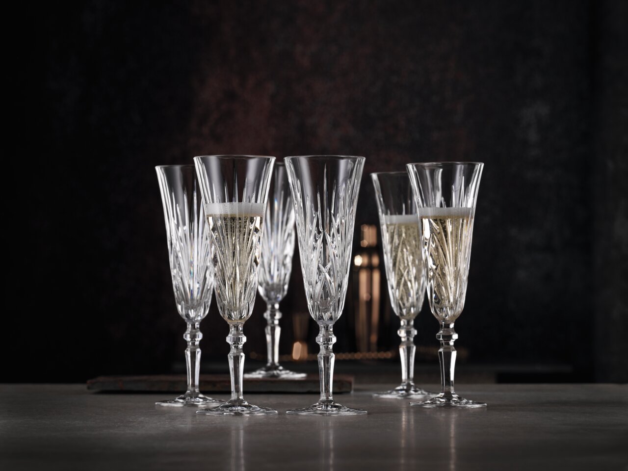 Sechs elegante Sektgläser aus Kristall mit feinem Schliff, teilweise gefüllt mit Sekt, auf einem Tisch vor dunklem Hintergrund, seitliche Perspektive.