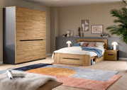 Modernes Schlafzimmer mit Holzbett inklusive Bettkasten und passendem Kleiderschrank in Eiche-Optik, Nachttisch und Lampen, fotografiert aus schräger Frontperspektive.