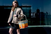 Zijaanzicht van een vrouw die een grote draagtas met een oranje kleurverloop en een lichte handgreep draagt, voor een moderne glazen gevel.