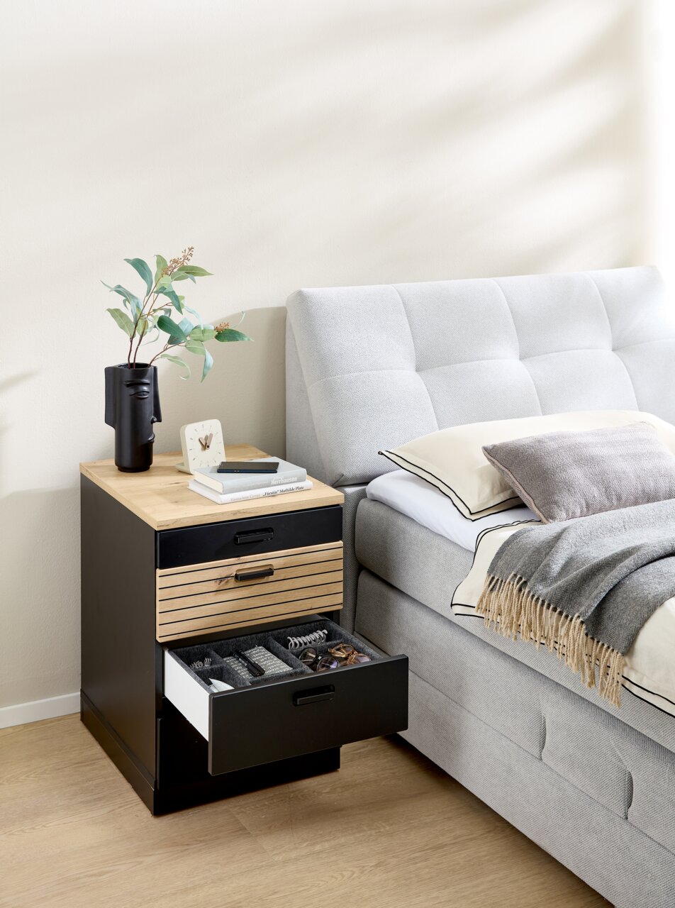 Rollcontainer mit drei Schubladen, aus Holz und Metall, neben einem Bett platziert. Perspektive von vorne.