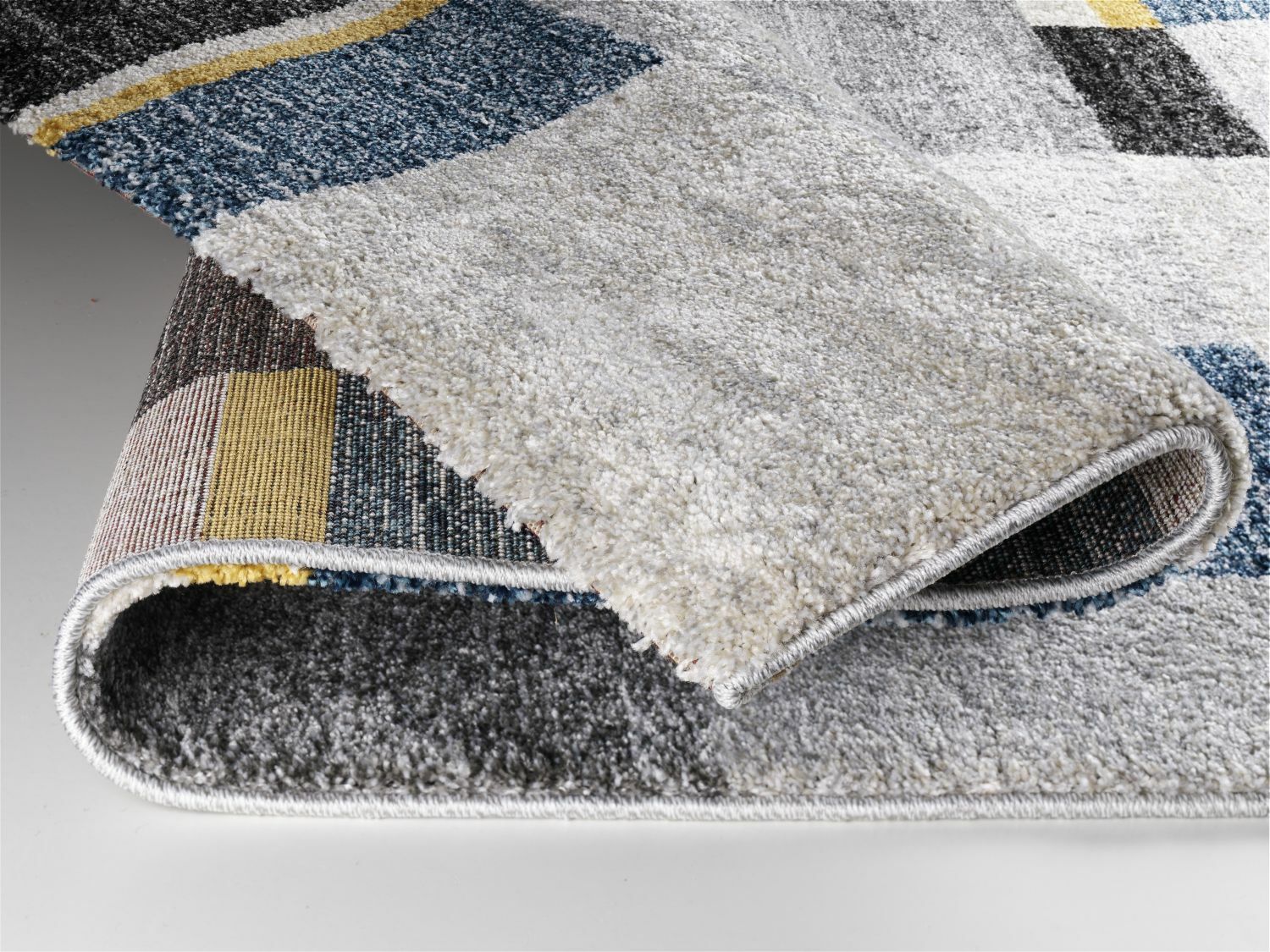 Nahaufnahme eines Teppichs mit geometrischem Muster in verschiedenen Farben, darunter Blau, Gelb und Grau. Der Teppich ist teilweise umgeschlagen, um die Dicke und Textur zu zeigen.