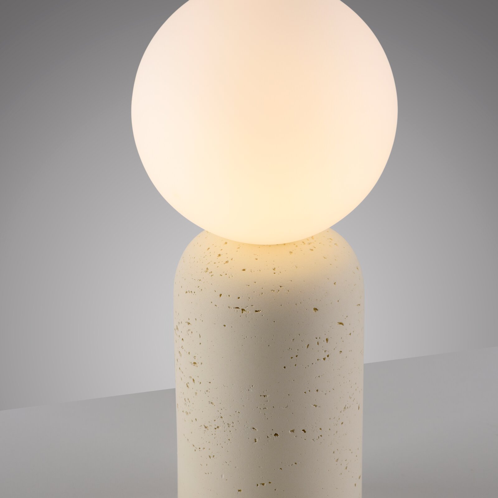 Minimalistische Tischlampe mit leuchtender Opalglaskugel auf zylindrischem, hellbeigem Steinfuß, frontale Nahaufnahme aus leicht erhöhter Perspektive.