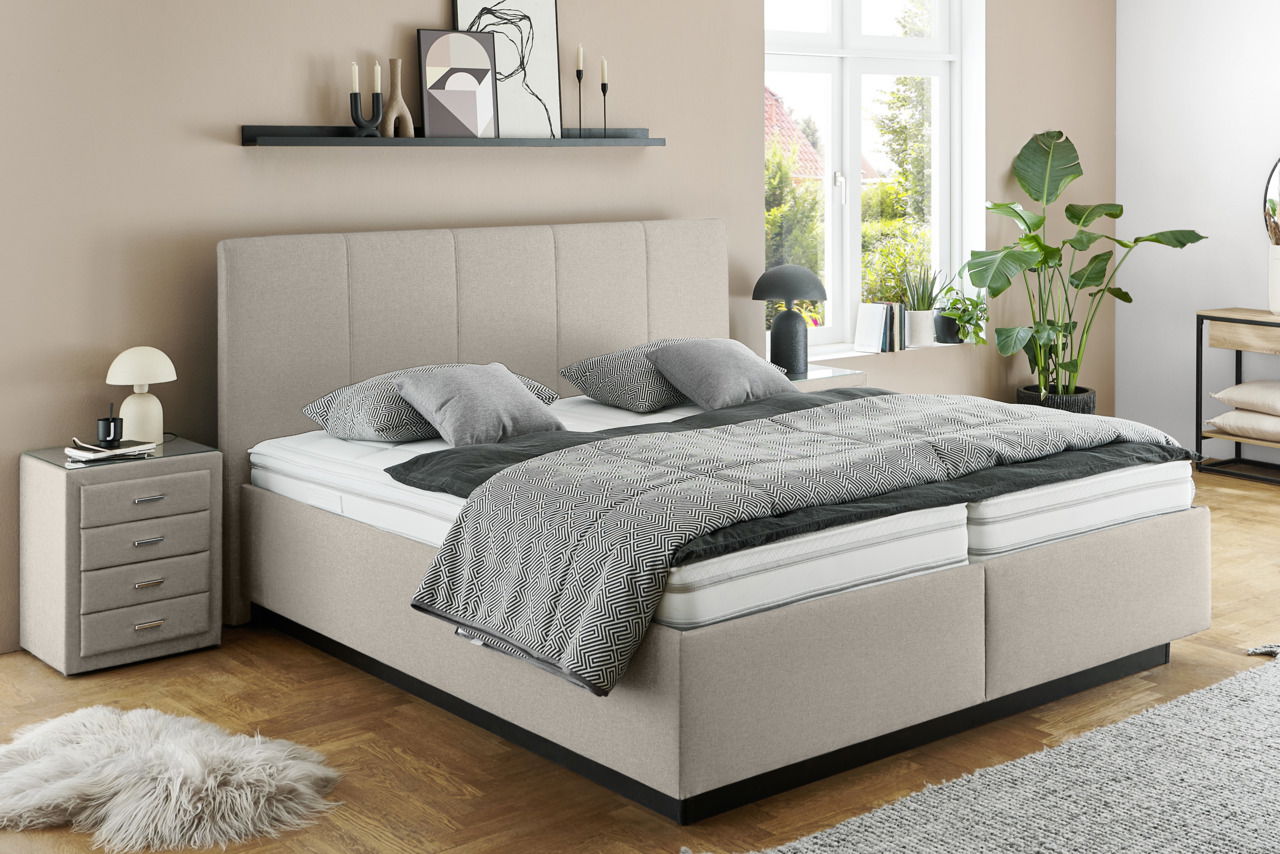 Modernes Polsterbett in Beige mit hohem Kopfteil, seitlicher Blick, dekoriert mit grauen Kissen und Decke, neben einem Nachttisch und Pflanzen.