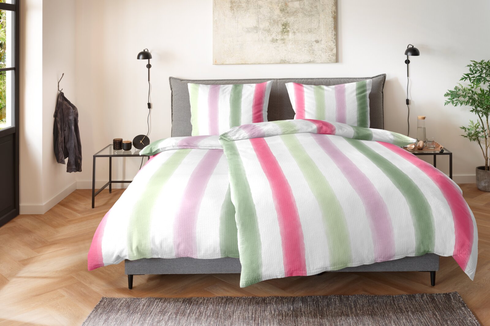 Gestreifte Bettwäsche in Weiß mit breiten Pastellstreifen in Grün, Rosa und Flieder auf einem grauen Polsterbett, frontal aus leichter Vogelperspektive im Schlafzimmer fotografiert.