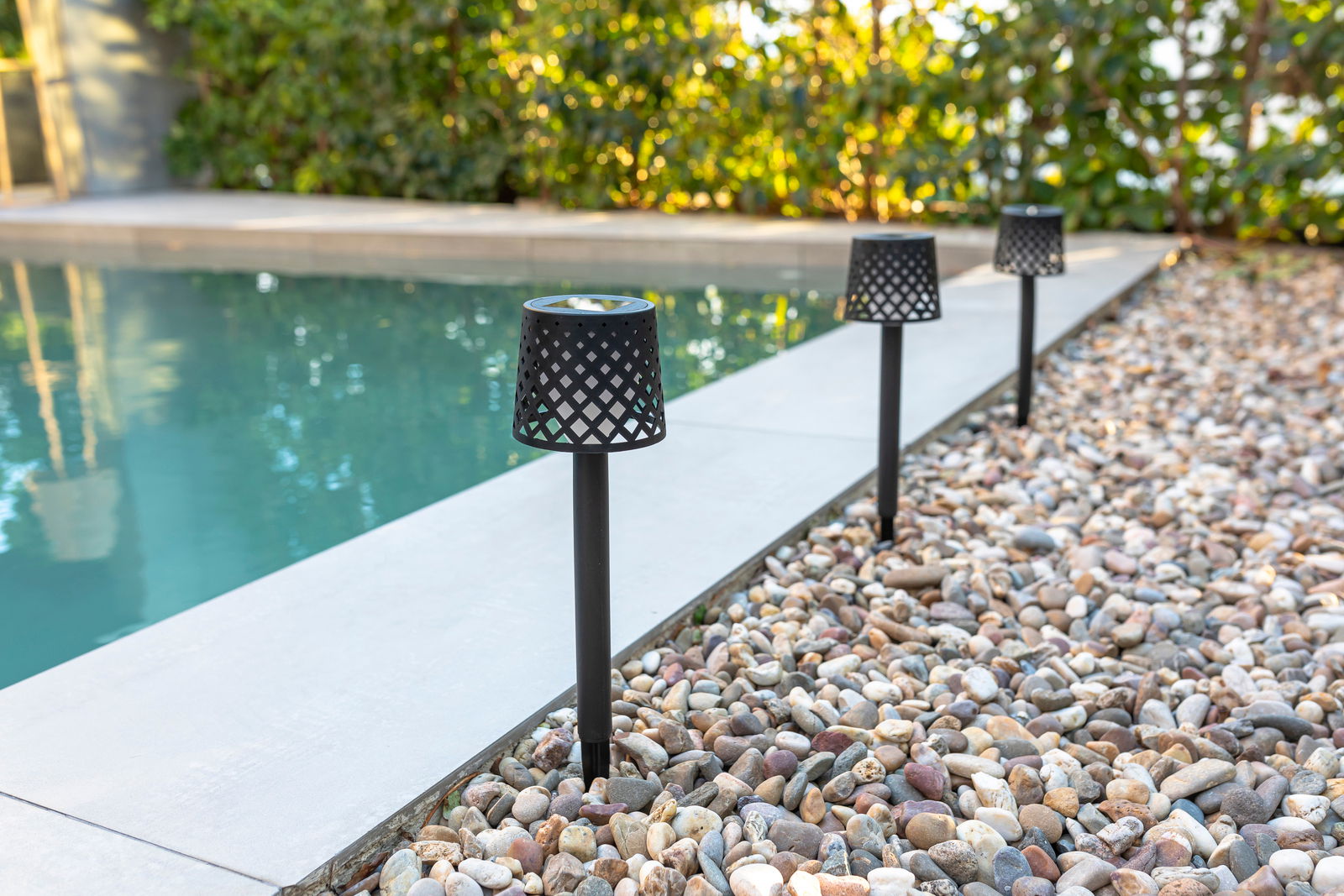 Drei schwarze Solar-Erdspieße mit Gitterdesign entlang eines Kiesweges neben einem Pool, seitliche Perspektive.