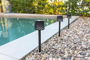 Drei schwarze Solar-Erdspieße mit Gitterdesign entlang eines Kiesweges neben einem Pool, seitliche Perspektive.
