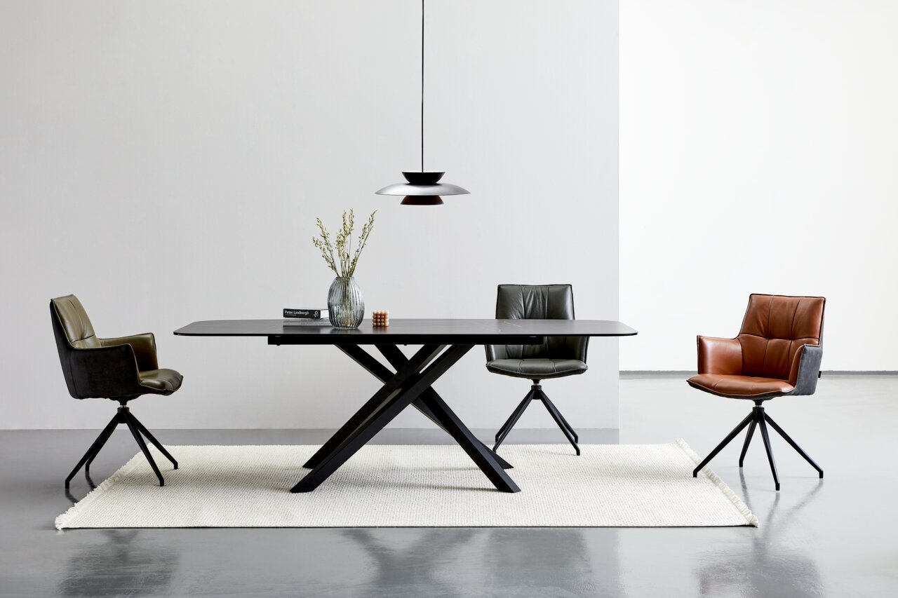 Moderner Esstisch mit schwarzer Tischplatte und X-förmigem Gestell, umgeben von drei stilvollen Stühlen in verschiedenen Farben, aus der Frontalperspektive.