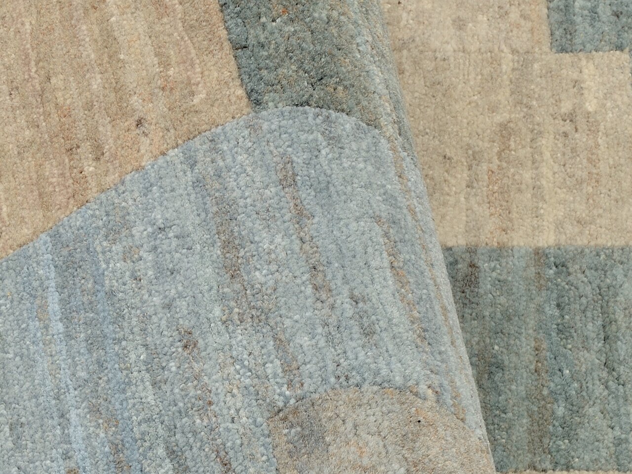 Nahaufnahme eines Wollteppichs mit geometrischem Muster in verschiedenen Beige- und Blautönen, teilweise aufgerollt.