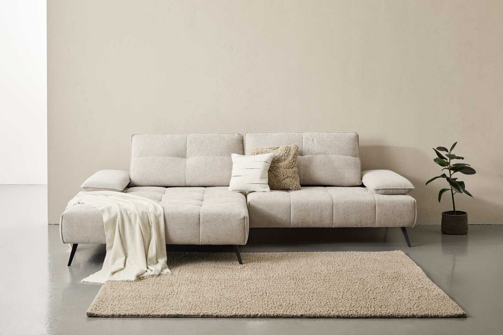 Beige Longchairkombination mit zwei Kissen, frontal fotografiert, auf einem Teppich stehend, mit einer Pflanze rechts daneben.