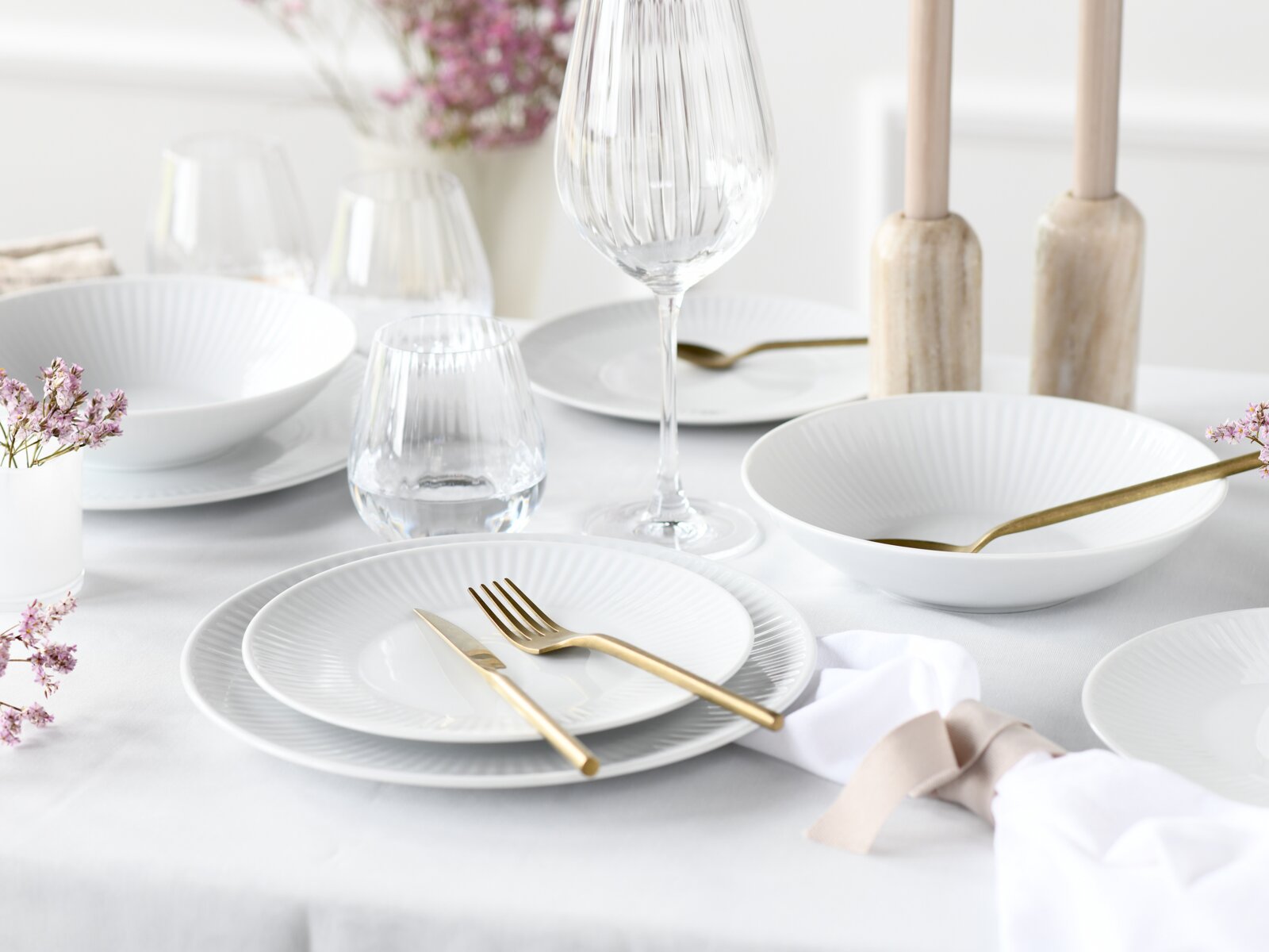 Elegant gedeckter Esstisch mit weißem geriffeltem Geschirr, goldfarbenem Besteck und Glaswaren, fotografiert aus leicht erhöhter, schräger Perspektive von vorne.