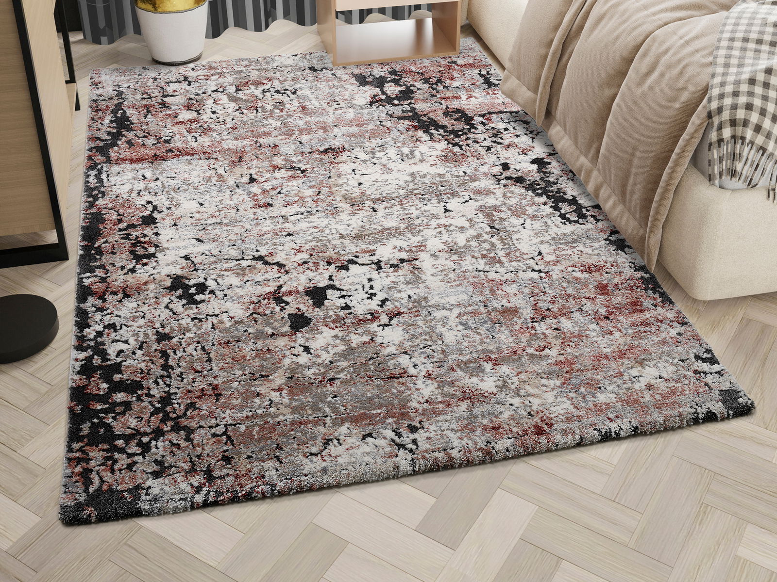 Designer Teppich mit abstraktem Muster in Rot-, Grau- und Schwarztönen, auf einem hellen Holzboden neben einem Bett, aus einer schrägen Perspektive fotografiert.