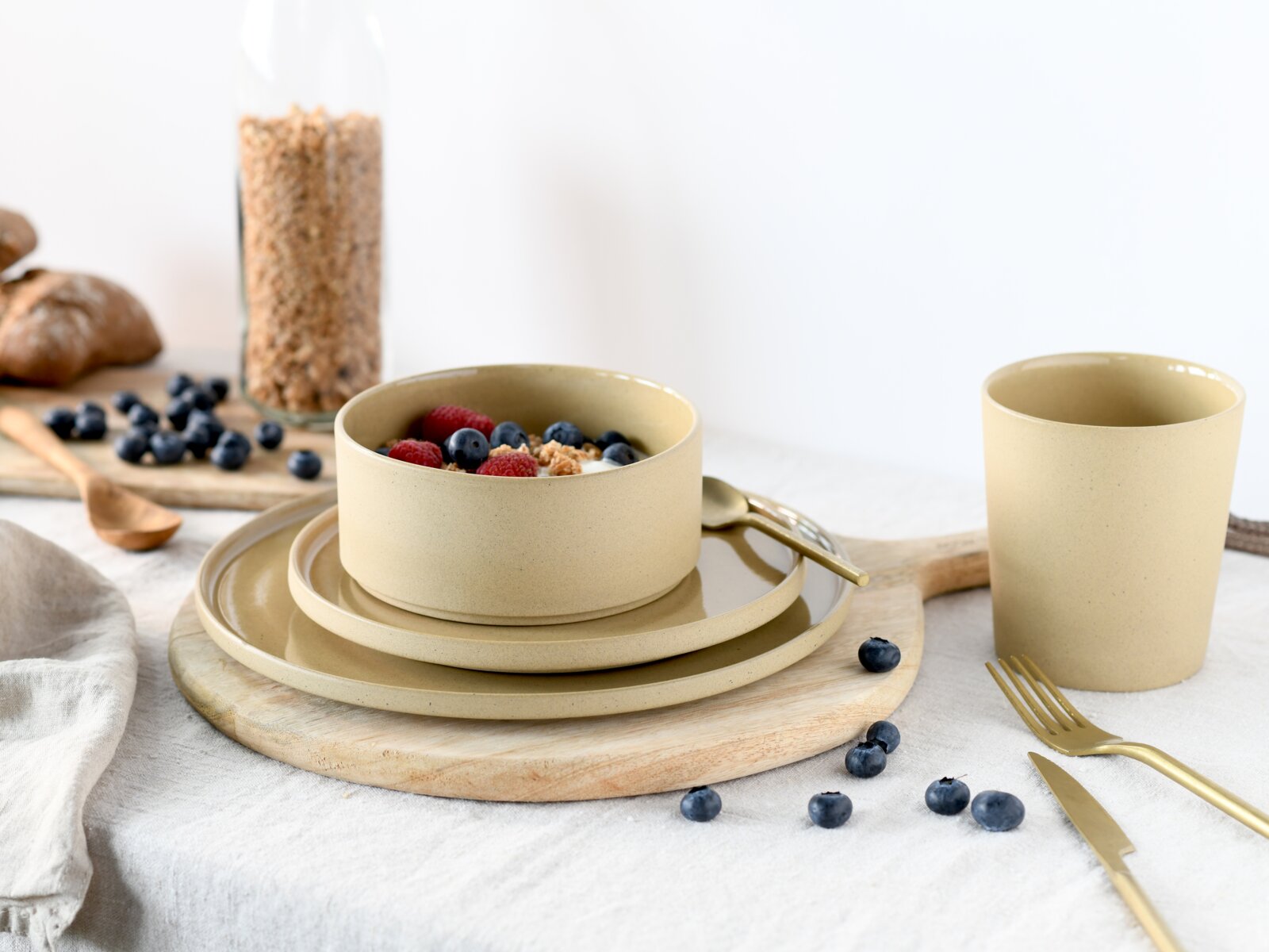 Beiges, mattes Keramik-Geschirrset mit Schüssel auf zwei Tellern und passendem Becher, arrangiert auf Holzbrett mit goldfarbenem Besteck und Beeren, fotografiert in leicht schräger Vorderansicht (3/4-Perspektive)