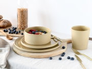 Beiges, mattes Keramik-Geschirrset mit Schüssel auf zwei Tellern und passendem Becher, arrangiert auf Holzbrett mit goldfarbenem Besteck und Beeren, fotografiert in leicht schräger Vorderansicht (3/4-Perspektive)