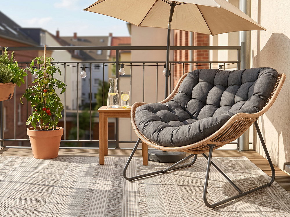 Gemütlicher Balkon mit gepolstertem Loungesessel, kleinem Holztisch, Sonnenschirm und bepflanzten Blumenkästen bei sonnigem Wetter.