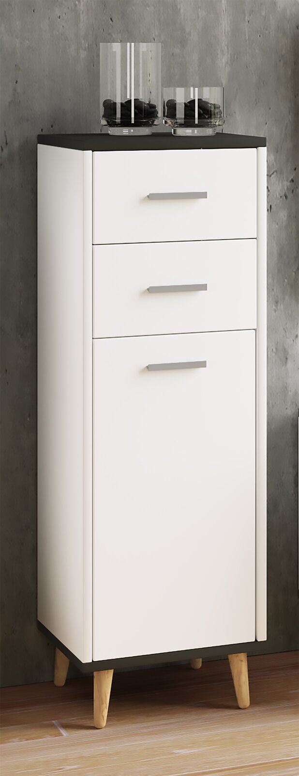 Weißer, schmaler Badezimmerschrank mit schwarzer Deckplatte, zwei Schubladen und Tür auf Holzfüßen, frontal in leichter Schrägansicht von vorn links fotografiert.