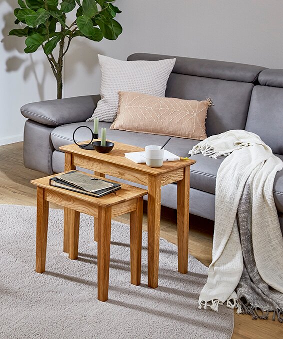 Zwei ineinanderschiebbare Beistelltische aus massivem Holz in natürlicher Eichenoptik vor grauem Sofa, dekoriert mit Kissen, Decke und Accessoires, fotografiert aus leicht erhöhter Frontperspektive.