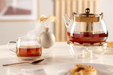 Glas-Teetasse mit Untertasse und dampfendem Tee, im Hintergrund eine Teekanne mit Teelicht, seitliche Perspektive