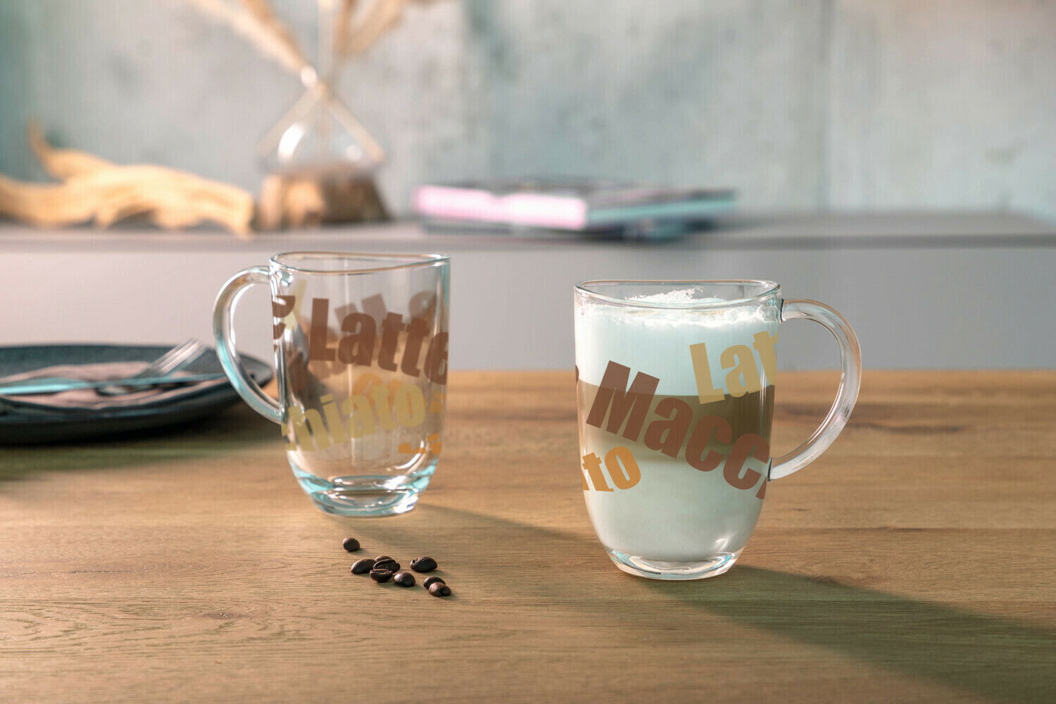 Zwei durchsichtige Glastassen mit Henkel, eine gefüllt mit Latte Macchiato, auf einem Holztisch. Im Hintergrund unscharf eine Vase und ein Teller. Perspektive: seitlich.